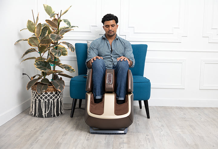 Man using the Zarifa Foot Massager+ while seated in a blue chair beside a houseplant